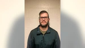 A man with short hair, a beard, and glasses stands against a light-colored cinder block wall, wearing a dark green jacket and a neutral expression—his look exudes the composed confidence often seen in marketing or public relations professionals.