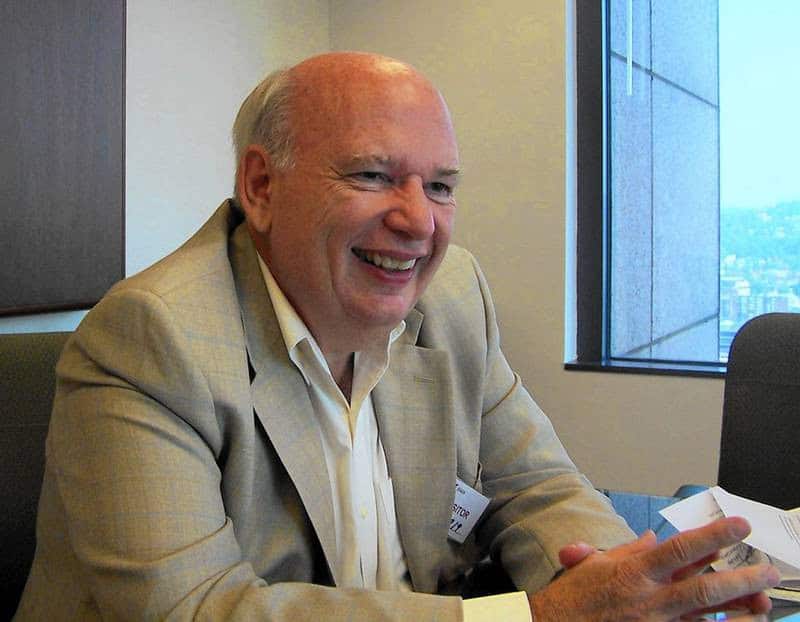An older man in a beige suit jacket and white shirt smiles while sitting at a table in an office, holding papers—perhaps preparing for a public relations meeting. City buildings are visible through the window in the background.