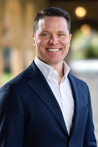 A smiling man in a navy blue suit and white shirt stands outdoors in a softly blurred walkway with warm lights in the background, capturing the polished confidence often seen in public relations and social media trends.