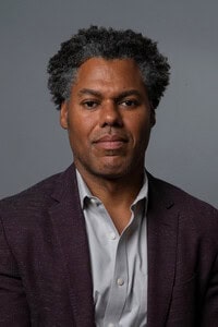 A man with short curly hair wearing a dark blazer over a light gray button-up shirt sits against a plain gray background, looking directly at the camera with a neutral expression, as if ready for media coverage or an interview in the news media.