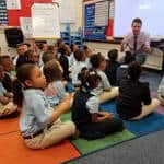 A teacher kneels at the front of a classroom, engaging a group of young students who are sitting cross-legged on a colorful rug, facing a projector screen displaying the SMART logo, discussing how social media trends can influence media coverage.
