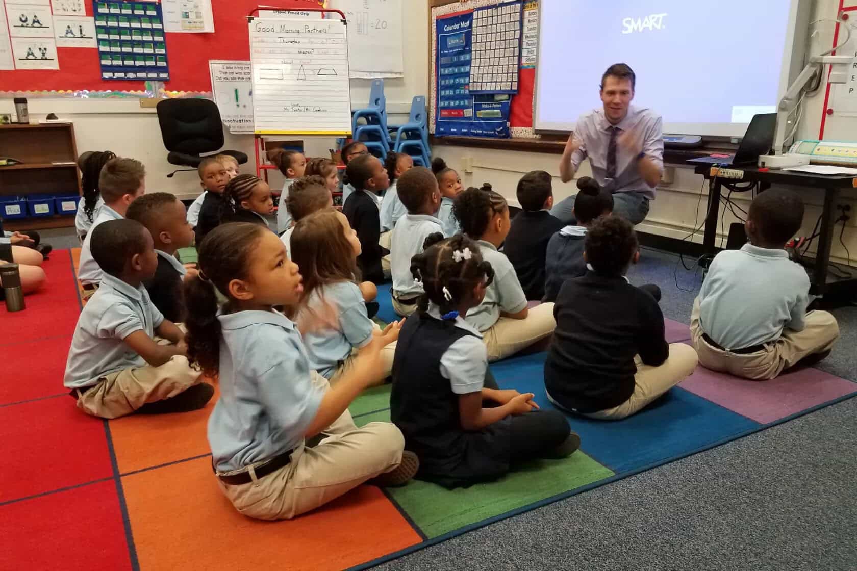 A teacher kneels at the front of a classroom, engaging a group of young students who are sitting cross-legged on a colorful rug, facing a projector screen displaying the SMART logo, discussing how social media trends can influence media coverage.
