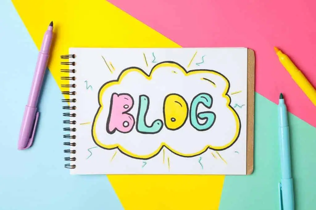 A spiral notebook with “BLOG” in colorful bubble letters sits on a pastel pink, blue, and yellow background—perfect for jotting down marketing ideas or tracking social media trends. Three uncapped markers—purple, yellow, and blue—surround the notebook.