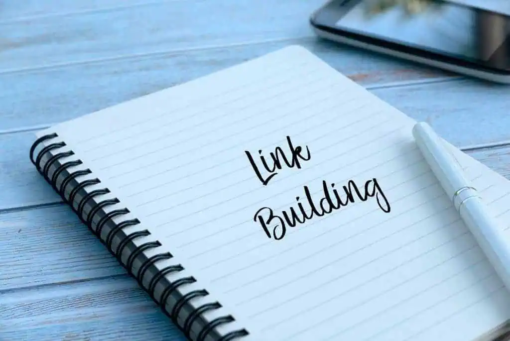 A notebook with the words Link Building written on a lined page sits beside a silver pen and smartphone, all placed on a light blue wooden surface—perfect for jotting down marketing ideas like Philly PR Girl or news media strategies.
