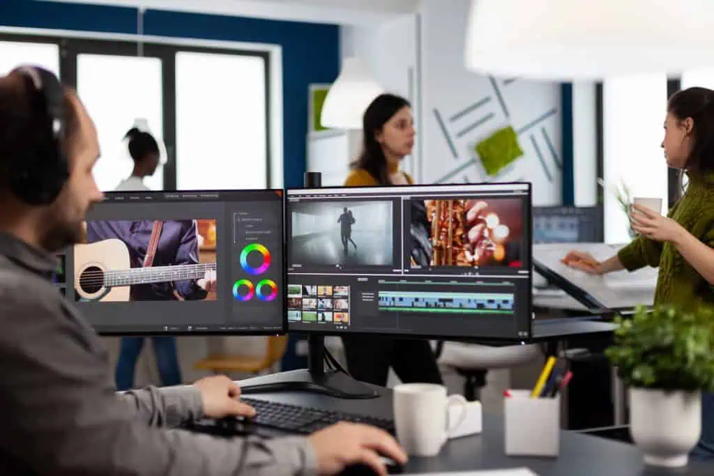 A man edits videos on dual monitors at a desk, incorporating social media trends and film clips. Three colleagues work and talk in the modern, brightly lit office background.
