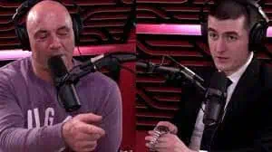 Two men wearing headphones sit in a recording studio with microphones, engaged in conversation about news media. One gestures as the other holds a metal object; red acoustic panels line the background, setting a professional broadcast scene.