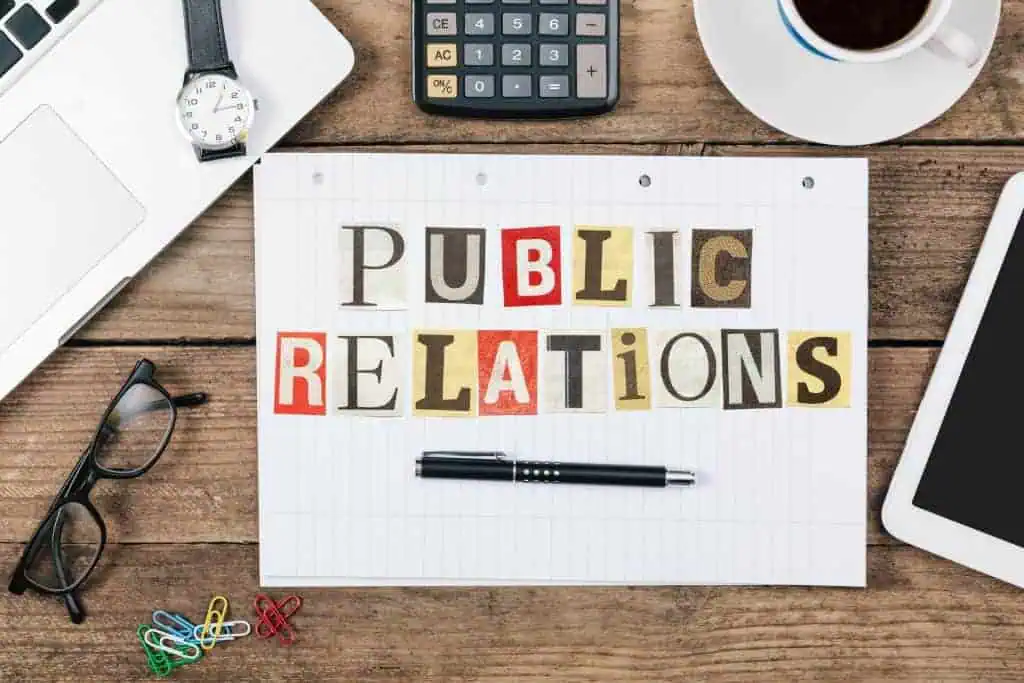 A sheet of lined paper with “Public Relations” spelled in colorful cut-out letters sits on a wooden desk, surrounded by a laptop, tablet, calculator, glasses, pen, coffee cup, and paper clips—perfect for marketing or tracking social media trends.