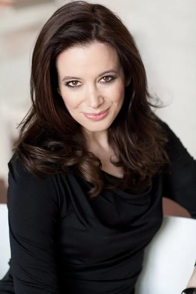 A woman with long, wavy brown hair and light skin is smiling softly at the camera. Dressed in a black top, she is seated against a light, blurred background—a pose often seen in social media trends.