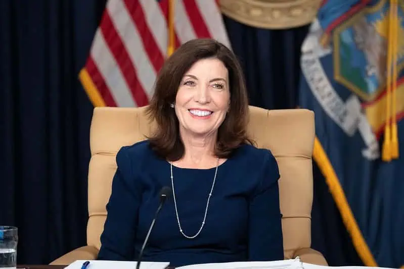 A woman with shoulder-length brown hair, wearing a navy blue dress and a necklace, sits and smiles at a desk with papers and a microphone—ready for her news media role. U.S. flags are visible in the background.