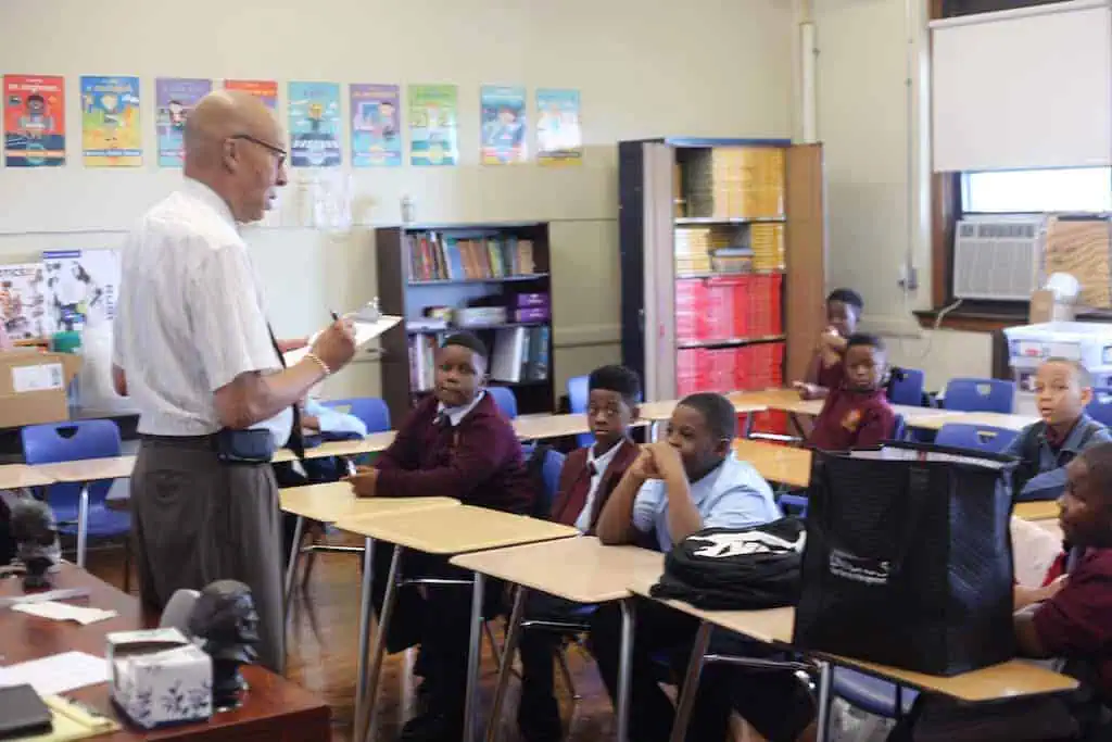 A teacher stands at the front of a classroom, holding a clipboard and speaking to attentive young students sitting at desks, as colorful posters and bookshelves highlight lessons about public relations and news media.