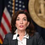 A woman with shoulder-length brown hair, wearing a white shirt and gray blazer, speaks at a podium with a U.S. flag and large gold seal in the background, addressing the news media in her role as a leading public relations professional.