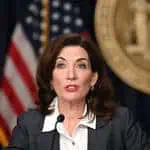 A woman with shoulder-length brown hair, wearing a white shirt and gray blazer, speaks at a podium with a U.S. flag and large gold seal in the background, addressing the news media in her role as a leading public relations professional.