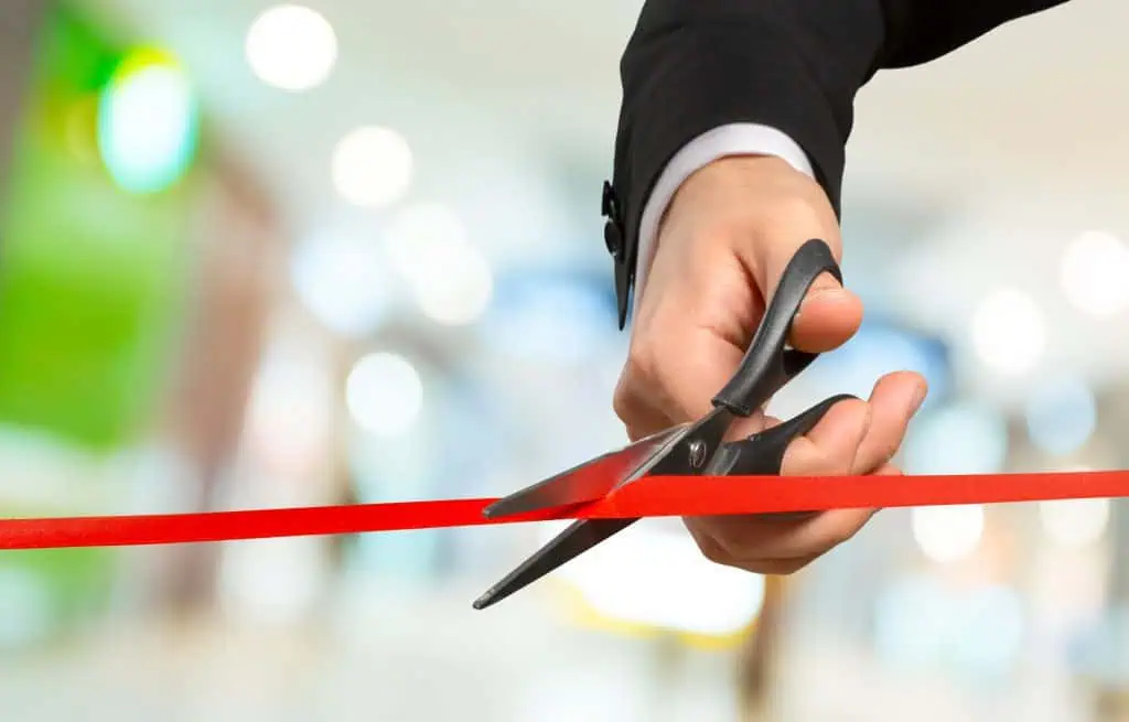 A person in a suit holds scissors and cuts a red ribbon, symbolizing the opening of an event or ceremony—an ideal moment for media coverage. The background is blurred with bright lights and indistinct shapes.