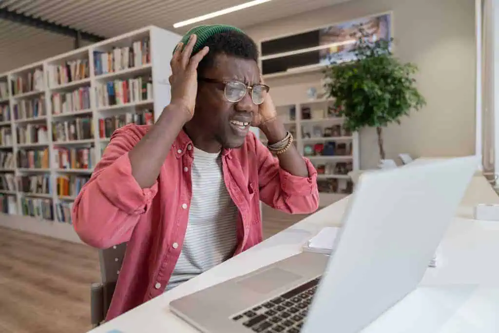 A young person wearing glasses and a green beanie looks frustrated while working on public relations tasks at their laptop in a library or study area. Bookshelves and a plant are visible in the background.