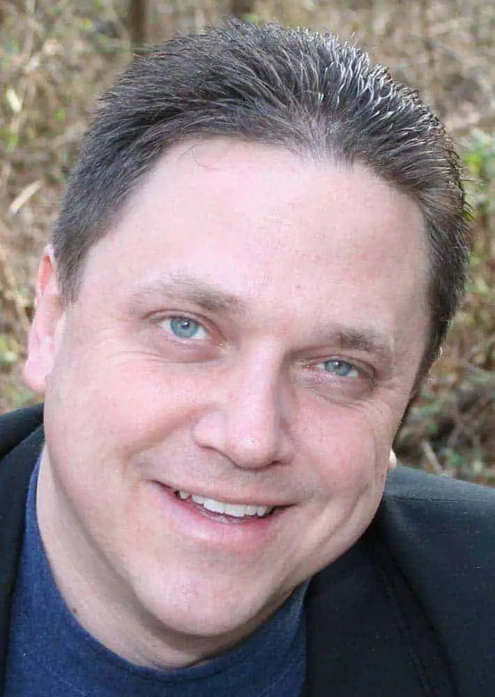 A man with short brown hair and blue eyes smiles at the camera. He is wearing a dark jacket and a blue shirt, posing outdoors with blurred brown foliage in the background, reflecting the confident style of a Philly PR Girl aware of social media trends.