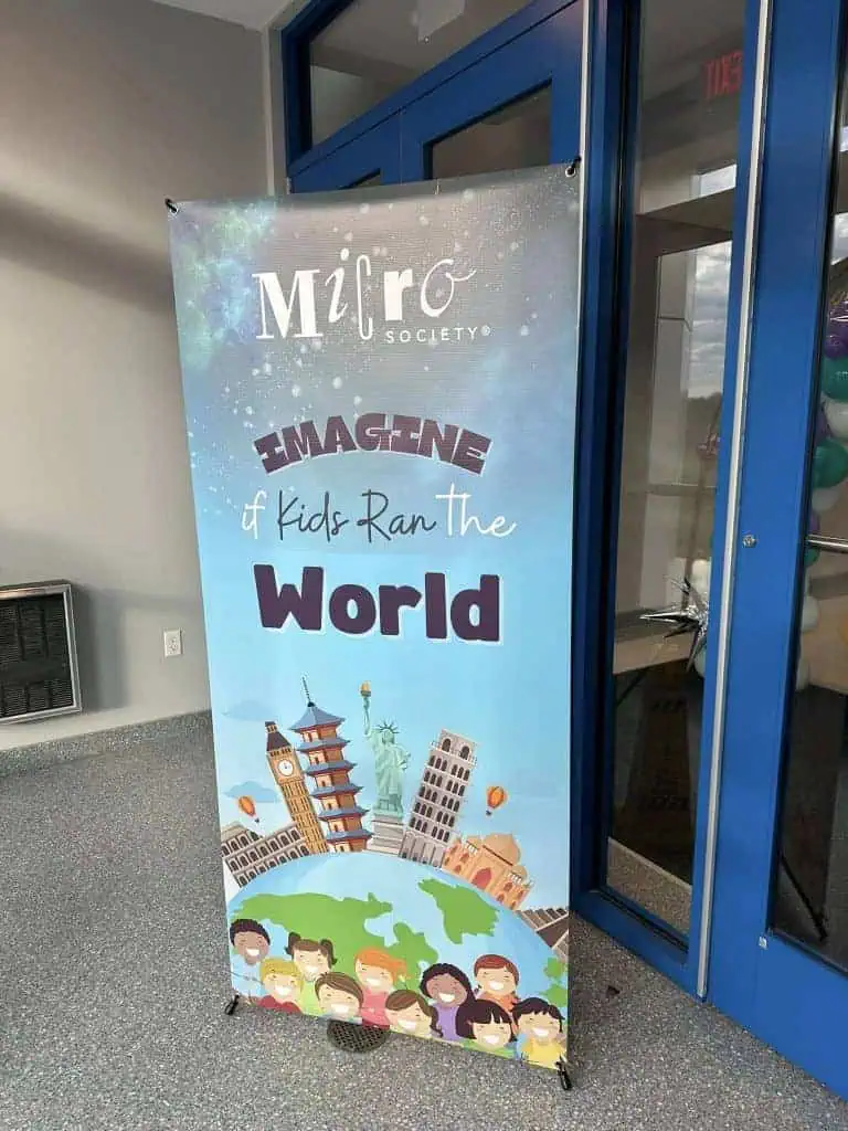 A colorful banner by a blue door reads “MiRo Society: Imagine if Kids Ran the World,” with illustrations of global landmarks and smiling children at the bottom—perfect for sharing on social media trends. Balloons are visible through the glass door.