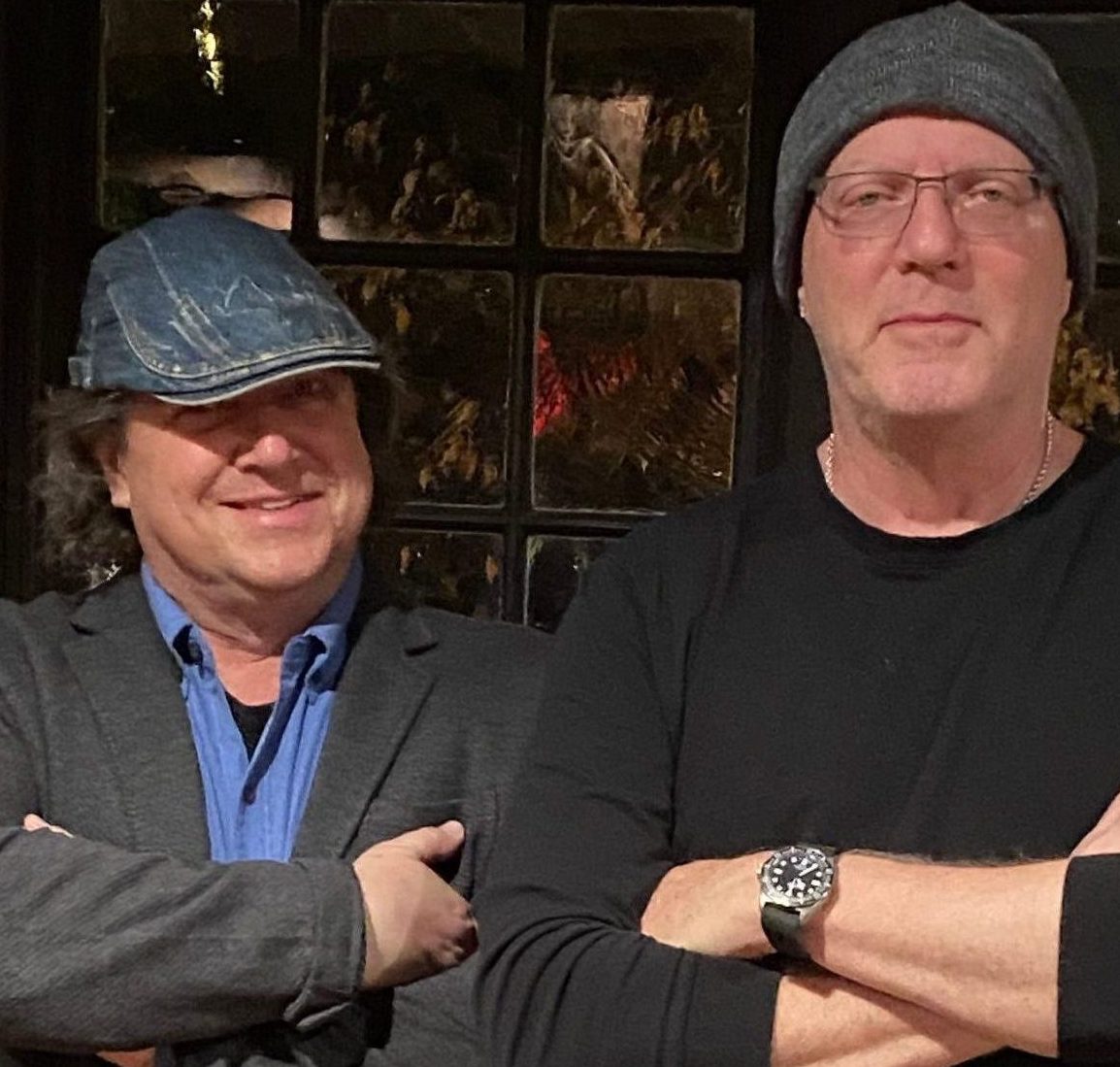 Two men stand side by side with arms crossed in front of a window, embodying the latest social media trends. One sports a blue denim cap and blazer; the other, glasses, a gray beanie, black shirt, and wristwatch.