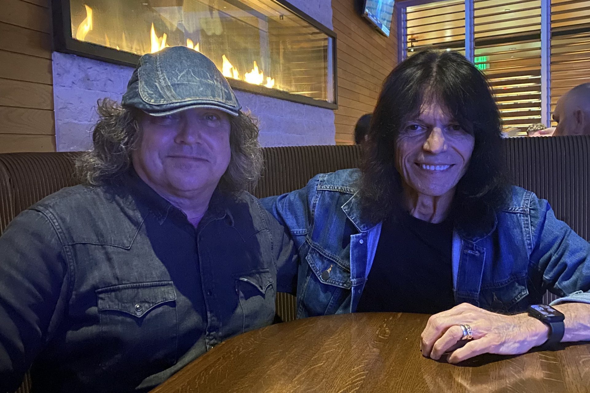 Rudy Sarzo and Ken Kilpatrick, both with long dark hair and denim shirts, sit together in a restaurant booth. A lit fireplace glows behind them while a wooden table sits in front. One man tops off his look with a denim cap.