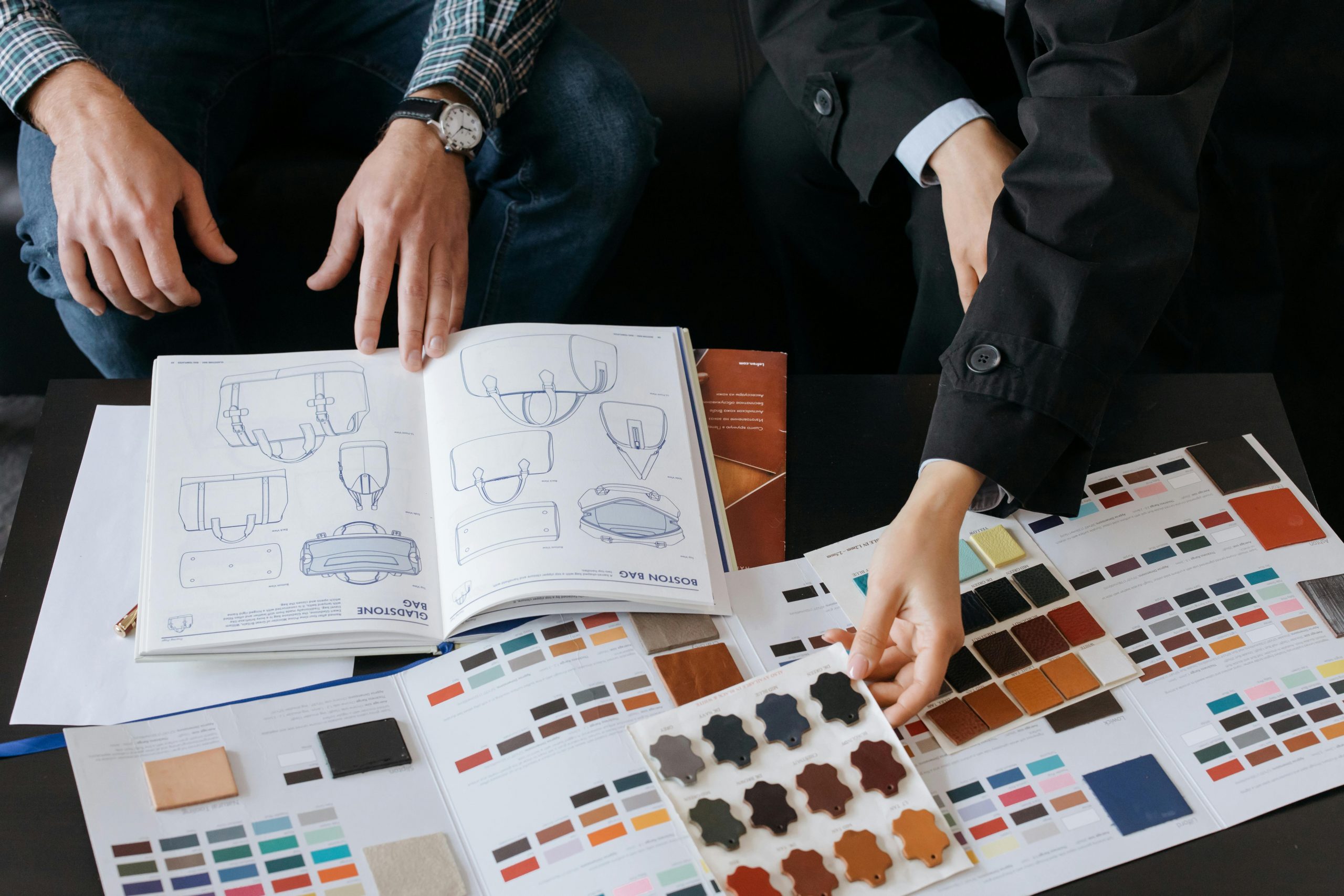 Two people sit at a table covered with color swatches, material samples, and open sketchbooks featuring technical car seat designs, discussing choices for upholstery and finishes.