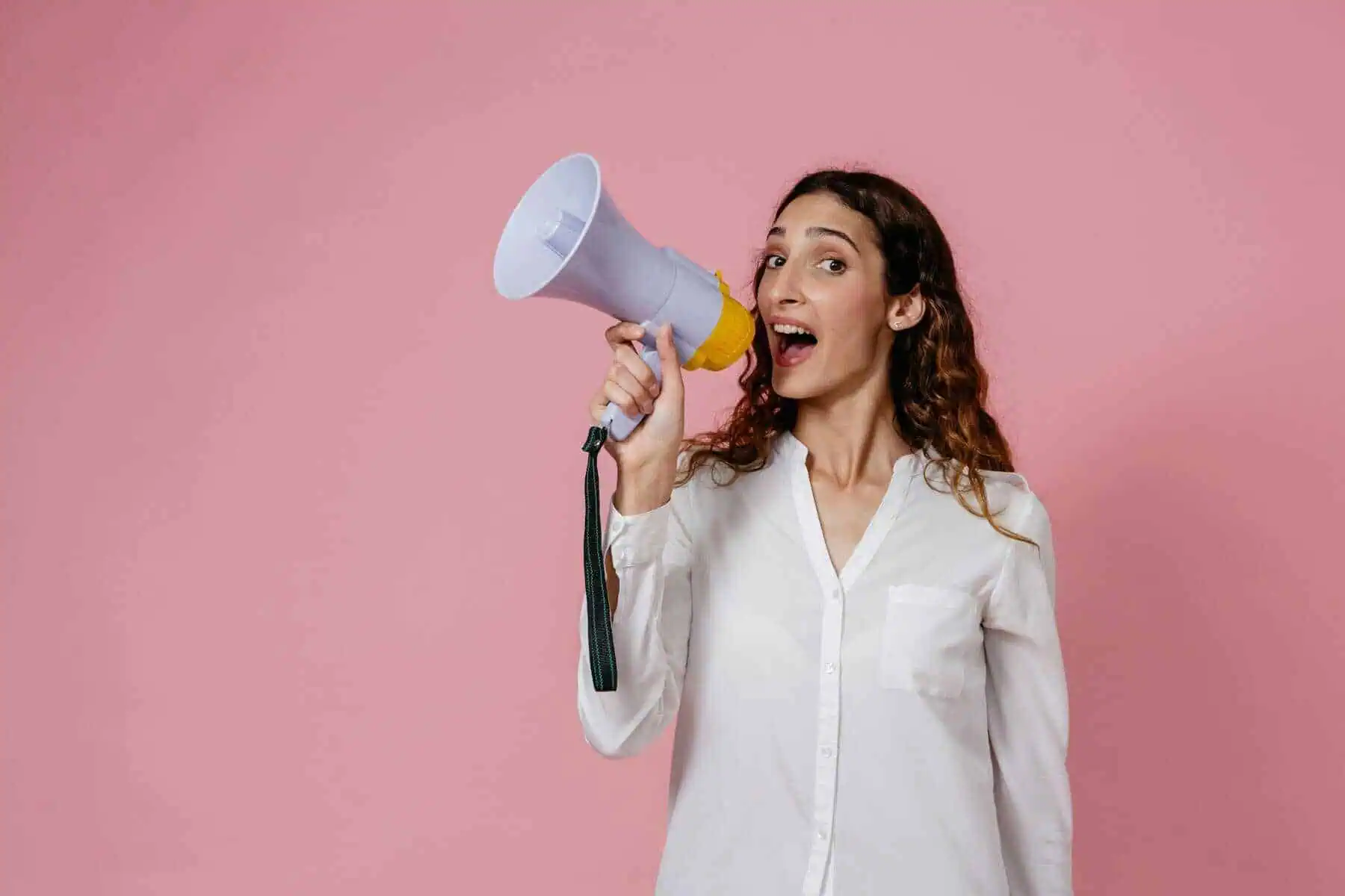 A woman with long curly hair, wearing a white shirt, smiles while holding a megaphone near her mouth against a pink background.