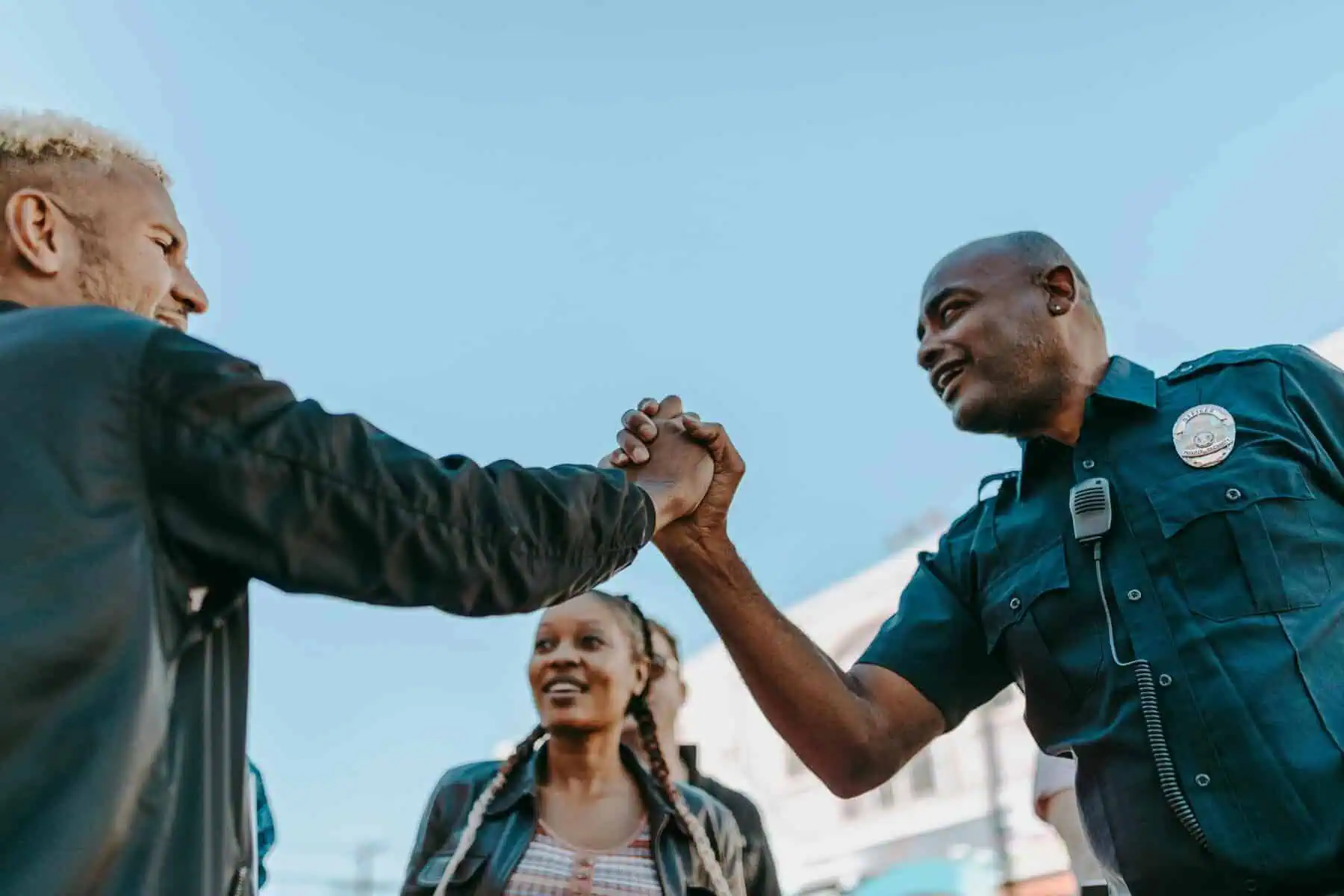 A police officer and a man clasp hands in a friendly handshake outdoors, smiling at each other. A woman stands between them, also smiling. The sky is clear and blue in the background.