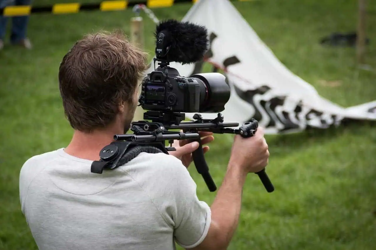A person with brown hair is filming outdoors with a professional camera mounted on a shoulder rig and microphone, standing on grass near a partially visible banner and black-and-yellow barrier.