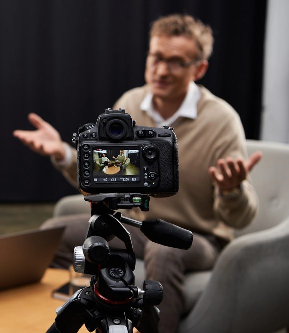 A camera on a tripod records two people sitting in chairs and talking. The man in the foreground gestures with his hands, while the woman listens. A laptop and a glass of water are on the table between them.