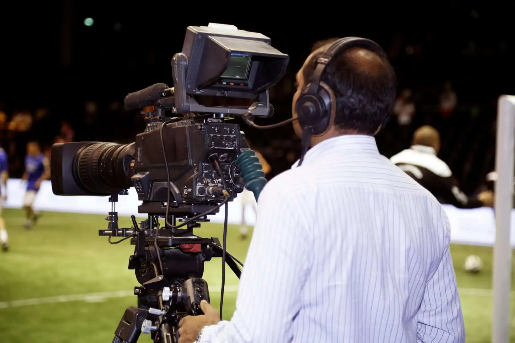 A camera operator wearing headphones films a soccer match from the sidelines, focusing on the field. Players and a coach are visible in the blurred background.